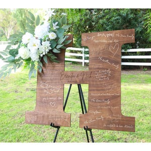 May include: A wooden letter H with a rustic finish, standing on an easel. The letter is decorated with a floral arrangement and personalized messages, including the names "Brenda & Nick" and the date "11.12.2023".