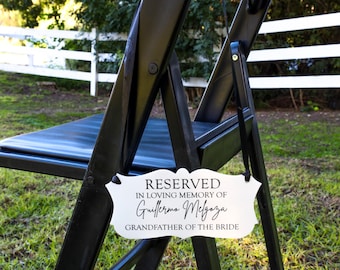 May include: A white sign with black text that reads "RESERVED IN LOVING MEMORY OF Guillermo Melgaza GRANDFATHER OF THE BRIDE" is attached to a black folding chair.
