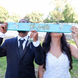 May include: A bride and groom hold a teal blue sign with the text "The Smiths" written in gold lettering. The sign is held above their heads.