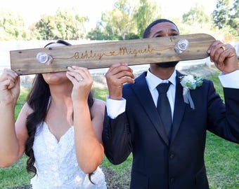 May include: A bride and groom hold a wooden sign with the text "Ashley & Miguel" in front of their faces. The sign has two small glass jars attached to it.