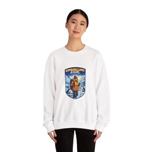 May include: White sweatshirt with a graphic featuring a hiker with the text "EXPLORE LIKE A GIRL". The graphic is in a shield shape with a snowy mountain scene and penguins. The hiker is wearing an orange jacket and a backpack.