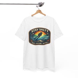 Puede incluir: Camiseta blanca con un gráfico colorido que presenta a un buceador en el océano bajo una puesta de sol. El diseño incluye el texto "STAY SALTY" arriba y "DIVE - EXPLORE - REPEAT" abajo. La camiseta está colgada en una percha de madera.