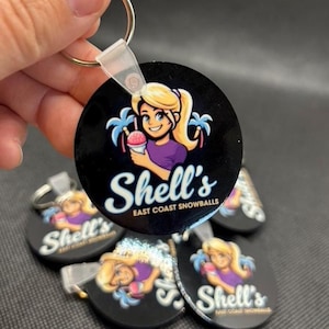 Könnte beinhalten: Ein schwarzer, runder Schlüsselanhänger mit einer Cartoon-Frau, die einen Schneekegel hält, mit dem Text "Shell's East Coast Snowballs". Die Frau hat blondes Haar und trägt ein lila Oberteil. Mehrere Schlüsselanhänger sind sichtbar.