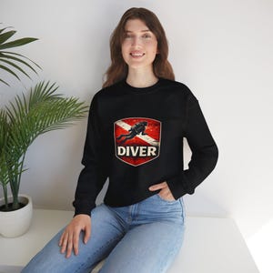 May include: Black sweatshirt featuring a red, white, and black graphic of a scuba diver inside a shield with the word "DIVER" below. The sweatshirt is a crew neck style.