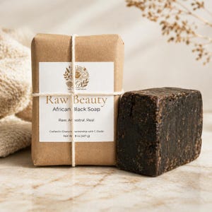 Puede incluir: Una barra de jabón negro africano marrón oscuro junto a una barra envuelta. La barra envuelta está en papel marrón con una etiqueta blanca que dice "Raw Beauty African Black Soap". El jabón está atado con hilo.
