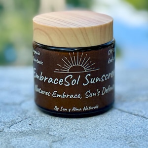 May include: A brown glass jar with a wooden lid, labeled "EmbraceSol Sunscreen" with the tagline "Natures Embrace, Sun's Defense" and the brand name "By Sun y Alma Naturals".