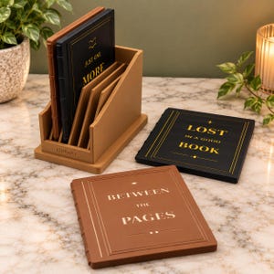 May include: A tan wooden organizer with the word LIBRARY embossed on the front, holding several square coasters designed like books. Two additional book-shaped coasters lie flat, one black with gold text reading LOST IN A GOOD BOOK and one tan with gold text reading BETWEEN THE PAGES.