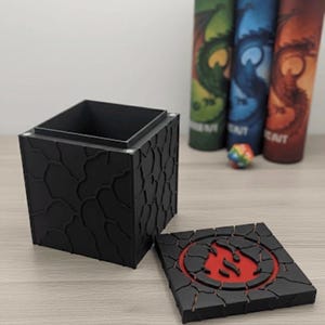 May include: A black, square box with a textured, cracked pattern. The lid is off, revealing a red flame design. In the background are three cylindrical containers with dragon illustrations and a colorful die.