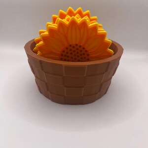 May include: A set of sunflower-shaped coasters in a brown basket-style holder. The coasters are yellow and orange, with a detailed petal design. The holder is a dark brown colour, with a woven basket design.