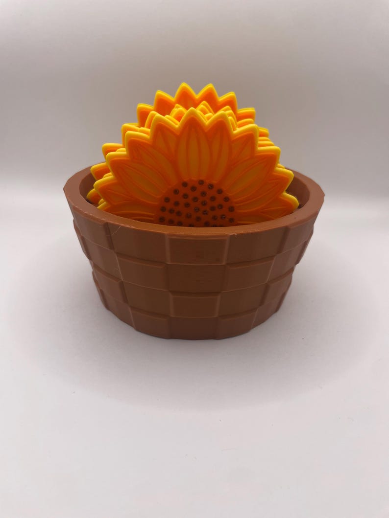 May include: A set of sunflower-shaped coasters in a brown basket-style holder. The coasters are yellow and orange, with a detailed petal design. The holder is a dark brown colour, with a woven basket design.