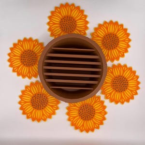 May include: A brown, circular, slotted container surrounded by six yellow and orange sunflower coasters. The coasters have a detailed design with a dark orange centre and yellow petals. The container is made of a solid brown material.