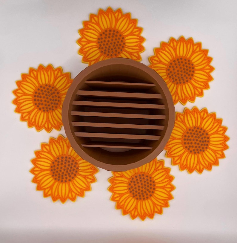 May include: A brown, circular, slotted container surrounded by six yellow and orange sunflower coasters. The coasters have a detailed design with a dark orange centre and yellow petals. The container is made of a solid brown material.