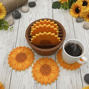 May include: Sunflower-shaped coasters in a brown holder, with a white mug of coffee on one. The coasters are yellow and orange, resembling sunflowers. The image is on a light wood-grain surface, with decorative sunflowers.