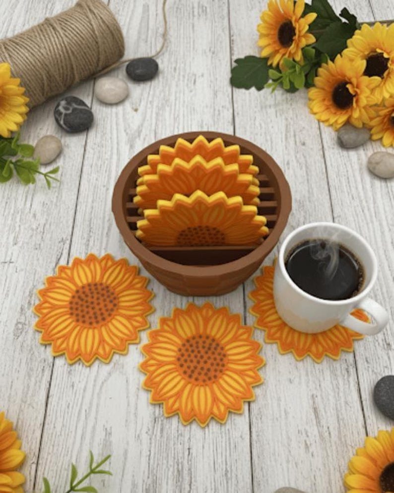 May include: A set of sunflower-shaped coasters, with a mug of coffee on one. The coasters are yellow and orange, and are stored in a brown holder. The image is on a light wood-grain surface, with decorative sunflowers.