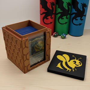 May include: A brown, hexagonal card storage box with a clear front panel, holding blue cards. A black lid with a yellow bee graphic sits beside it. Three cylindrical containers with dragon silhouettes in red, green, and blue are in the background.