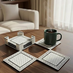 May include: A set of four square coasters with a matching holder. The coasters have a white background with a black geometric pattern and a black border. A green mug sits on one coaster. The holder is white with black accents.
