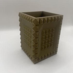May include: A square, olive-green container with a textured, pyramid-like pattern. The container has a hollow interior and is likely used for storage or display. The design features repeating geometric shapes, creating a modern aesthetic.