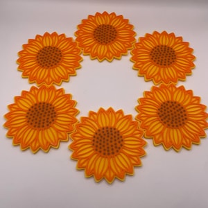 May include: Six sunflower-shaped coasters. Each coaster has a bright yellow and orange design, with a detailed centre and petal pattern. The coasters are arranged in a circular pattern, ideal for adding a touch of summer to any space.