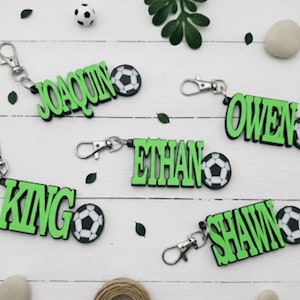 May include: Green name keychains with soccer ball accents. The names "Joaquin", "Owen", "Ethan", "King", and "Shawn" are displayed. Each keychain has a silver clasp. Decorative elements include small soccer balls, leaves, and stones.