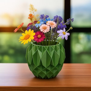 May include: A vibrant floral arrangement in a geometric green vase. The bouquet features a variety of colorful flowers, including yellow, pink, and blue blooms. The vase is made of a dark green material and has a faceted design. The arrangement sits on a wooden surface.