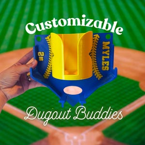 May include: A blue and yellow baseball-themed holder with the text "Customizable" and "Dugout Buddies". The holder has a yellow interior and blue exterior with the name "MYLES" and the number "2" on the sides. The background is a baseball field.