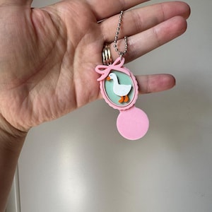 Puede incluir: Un llavero con forma de medallón ovalado de color rosa que presenta un fondo verde claro con una ilustración de un ganso blanco. El medallón tiene un pequeño lazo rosa en la parte superior y una cadena de bolas plateada. La parte inferior es un disco rosa.