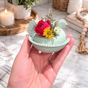 Könnte beinhalten: Hellgrüne Keramik-Henne-förmige Schmuckschale, gefüllt mit einem lebendigen Arrangement aus roten, gelben und weißen Blumen. Die Schale wird in einer Hand gehalten, die ihr detailliertes Design und die dekorative Blumenanordnung zeigt. Der Hintergrund umfasst eine brennende Kerze und andere Wohnaccessoires.