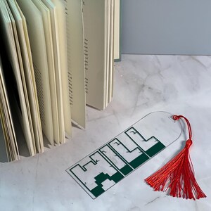May include: A clear glass bookmark with a green pixelated design of the word "HILL" and a red tassel.