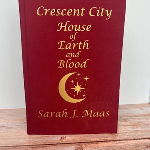 May include: A red hardcover book with the title "Crescent City House of Earth and Blood" by Sarah J. Maas. The cover features a gold crescent moon with a star.