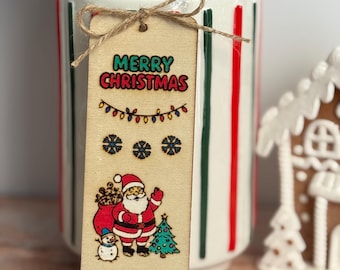 May include: A wooden Christmas bookmark with a festive design. The bookmark features the text "MERRY CHRISTMAS" and "Ho Ho Ho", along with illustrations of Santa Claus, a snowman, and a Christmas tree. The bookmark is tied with twine.