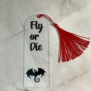 May include: A clear acrylic bookmark with a black dragon silhouette and the text "Fly or Die". The bookmark has a red tassel.
