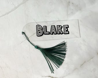 Puede incluir: Un marcapáginas de acrílico transparente con el nombre "BLAKE" impreso en letras negras. El marcapáginas tiene un borla verde.