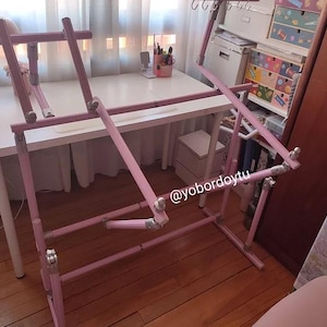 May include: A pink adjustable craft stand, likely for cross-stitch or embroidery. The stand is made of pink metal tubing and has adjustable angles and heights. The stand is set up in a well-lit room with a white desk and other craft supplies.