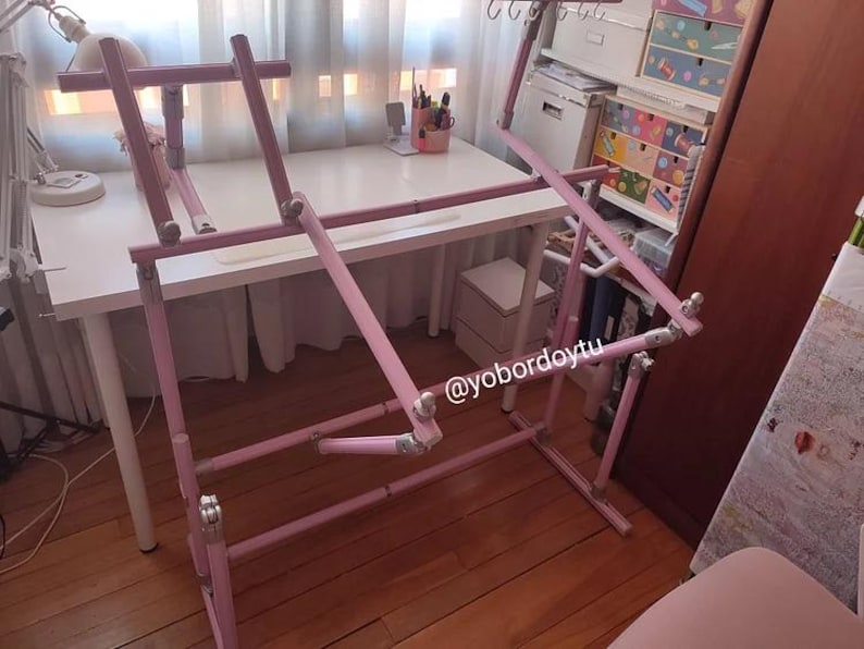 May include: A pink adjustable craft stand, likely for cross-stitch or embroidery. The stand is made of pink metal tubing and has adjustable angles and heights. The stand is set up in a well-lit room with a white desk and other craft supplies.