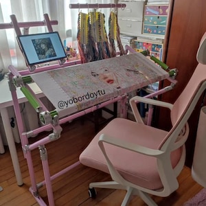May include: A pink cross-stitch frame with a completed embroidery project, a tablet, and a pink office chair. The frame is adjustable and has green accents. A variety of embroidery floss hangs above the frame. The tablet displays an image of a woman.