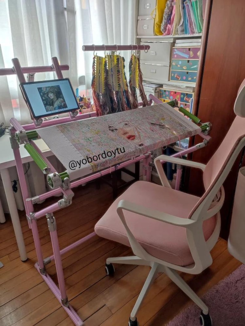 May include: A pink cross-stitch frame with a completed embroidery project, a tablet, and a pink office chair. The frame is adjustable and has green accents. A variety of embroidery floss hangs above the frame. The tablet displays an image of a woman.
