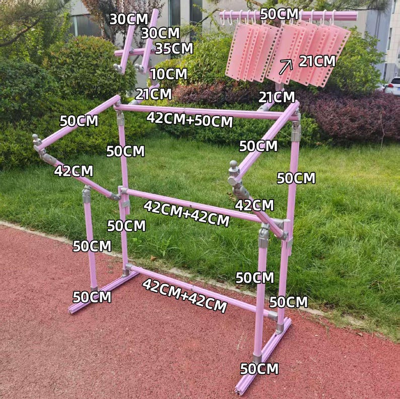 May include: A pink metal clothes drying rack with multiple horizontal bars and vertical supports. The rack has measurements labeled in centimeters, including 50 cm, 42 cm, and 30 cm sections. Several pink plastic hangers are attached to the top.