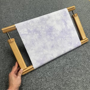 May include: A wooden embroidery frame holding a piece of fabric. The fabric is white with a light purple tie-dye pattern. The frame is made of light-coloured wood and has adjustable screws. The fabric is approximately 30 cm wide.