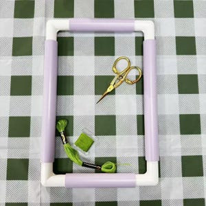 May include: A rectangular embroidery hoop with lavender-colored sides and white corners, accompanied by gold scissors, green embroidery floss, and a small square of green fabric. The items are arranged on a green and white checkered surface.