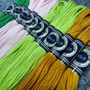 May include: A collection of embroidery floss in various colors, including green, pink, yellow, and gold. Each skein is wrapped with a blue and white label that reads "Cross Stitch" and "8.7 yd". The floss is arranged in a diagonal pattern.