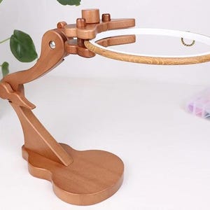 May include: A wooden embroidery hoop stand with a curved arm and a round base. The stand holds a white and wood-toned embroidery hoop. The stand is made of light brown wood and is designed to hold an embroidery project.