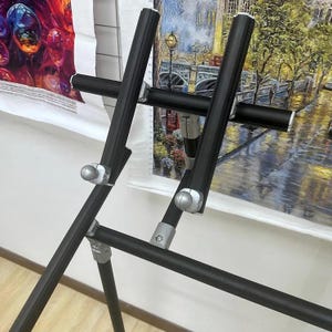 May include: A black metal easel with adjustable height and width, designed to hold artwork. The easel has a tripod base for stability and features silver-colored adjustment knobs. Two pieces of artwork are visible in the background.