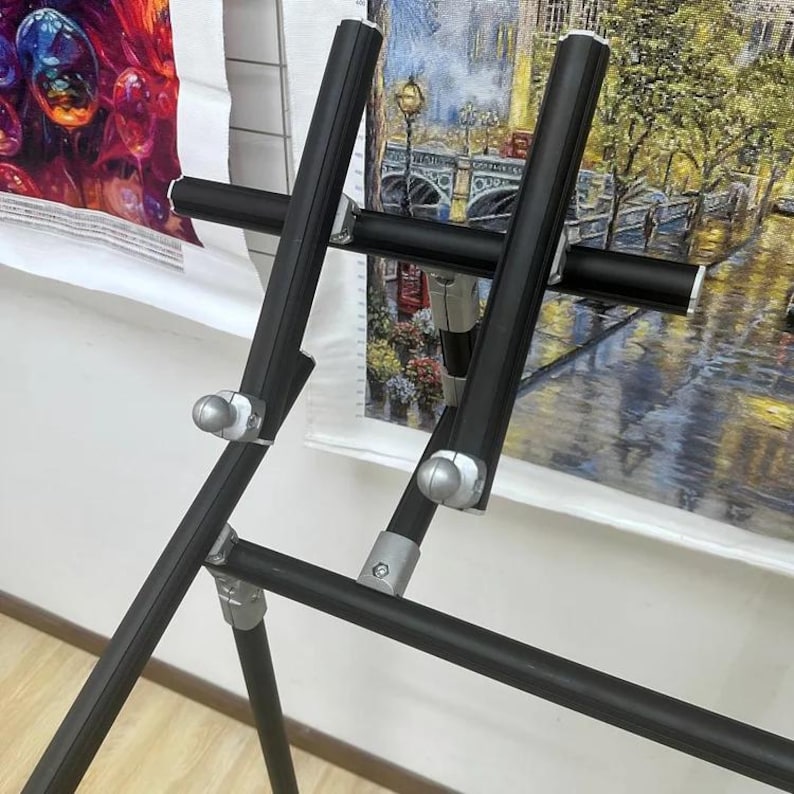 May include: A black metal easel with adjustable height and width, designed to hold artwork. The easel has a tripod base for stability and features silver-colored adjustment knobs. Two pieces of artwork are visible in the background.