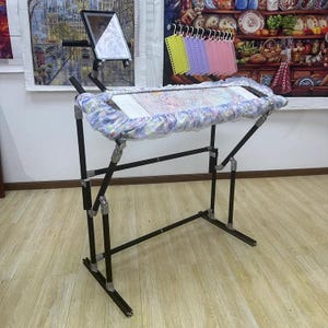 May include: A black metal embroidery stand with a fabric-covered frame. The stand has adjustable legs and a built-in tablet holder. The frame holds a piece of fabric with a colorful, detailed pattern. A set of colorful thread organizers hangs above the frame.