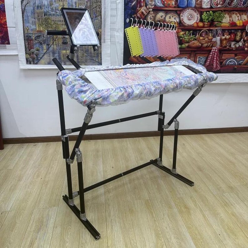 May include: A black metal embroidery stand with a fabric-covered frame. The stand has adjustable legs and a built-in tablet holder. The frame holds a piece of fabric with a colorful, detailed pattern. A set of colorful thread organizers hangs above the frame.