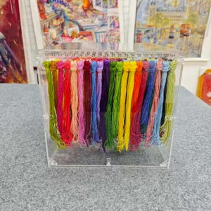 Op de afbeelding: Een doorzichtige acryl draadorganizer gevuld met kleurrijk borduurgaren. Het garen is gerangschikt in een regenboog van kleuren, waaronder groen, geel, oranje, rood, roze, paars en blauw. De organizer heeft een liniaal op de bovenrand.
