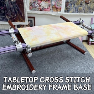 Peut inclure: Base de cadre de broderie au point de croix de table. Le cadre est composé d'un tissu de couleur claire tendu. Le cadre est en bois marron et des barres violettes avec des pinces réglables argentées. Le texte "TABLETOP CROSS STITCH EMBROIDERY FRAME BASE" est en bas.