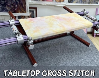May include: Tabletop cross stitch embroidery frame base. The frame has a light-colored fabric stretched across it. The frame is made of brown wood and purple bars with silver-colored adjustable clamps. The text "TABLETOP CROSS STITCH EMBROIDERY FRAME BASE" is at the bottom.