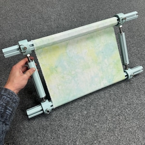 May include: A light blue adjustable embroidery hoop with a piece of fabric stretched across it. The fabric has a watercolor design in shades of green and yellow. The hoop is held by a person's hand.