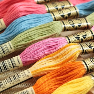 May include: Close-up of colorful embroidery floss spools in shades of pink, blue, green, yellow, and orange. Each spool has a label with text and is approximately 8.7 yards in length. The floss is tightly wound and ready for crafting projects.
